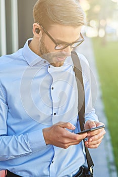 Making important call. Young handsome businessman wearing wireless earphones using smartphone while standing outdoors