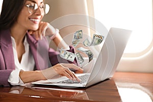 Making money online. Woman using laptop at table and flying dollars