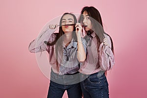 Making mustache with hair. Young women having fun in the studio with pink background. Adorable twins