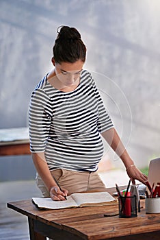 Making notes for when she comes back from maternity leave. a young pregnant woman writing in her diary while standing in