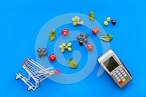 Making purchase online with card machine, mini trolley and products on blue desk background top view