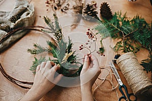 Making rustic christmas wreath. Hands holding berries, fir branches, pine cones, thread, scissors on wooden table.  Authentic