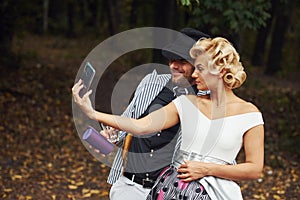 Making a selfie. Beautiful couple in old fashioned wear near retro car with forest at background