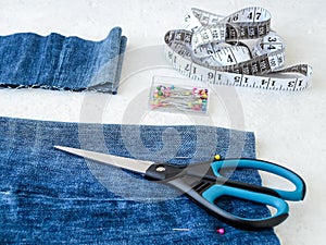 Making shorts out of blue jeans with multi colored headed sewing pins, white tailor tape with centimeters and inches and scissors