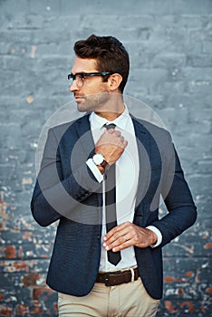 Making sure he looks his best. a handsome young businessman adjusting his tie while standing against a grey facebrick