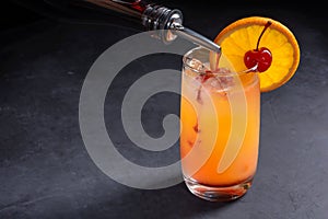 Making Tequila Sunrise Cocktail. Grenadine slowly poured into a glass of ice, orange juice and tequila. Dark stone background with