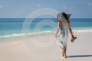 Maldives, young woman walking along the beach with sunhat and high heels on the hand