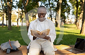 Male african american student reading book outdoors, preparing for classes, sitting in park or university campus