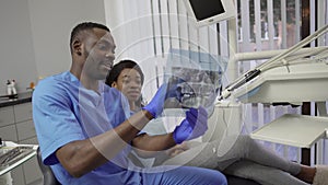 Male african dentist speaking with patient, pretty dark skinned girl, in dental clinic, showing her panoramic dental X