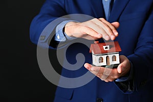 Male agent covering house model on black, closeup. Home insurance