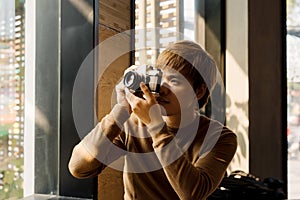 Male amateur photographer in eyeglasses focusing to make photos out of window sitting on windowsill.Young bearded man resting in