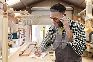 Male Apprentice Working As Carpenter In Furniture Workshop Making Phone Call
