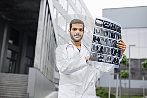 male Arabian Indian doctor, holding patient's tomography scan, posing near modern hospital