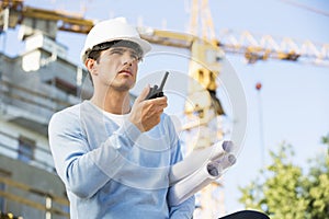 Male architect with blueprints using walkie-talkie at construction site