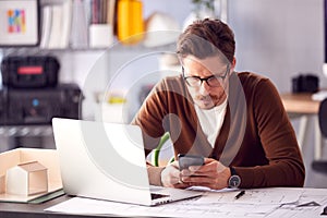 Male Architect In Office With Laptop Working On Plan At Desk Text Messaging On Mobile Phone