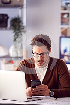 Male Architect In Office With Laptop Working On Plan At Desk Text Messaging On Mobile Phone