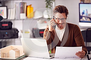 Male Architect In Office Working At Desk On Laptop Taking Call On Mobile Phone