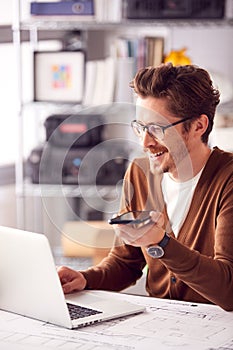 Male Architect In Office Working At Desk On Laptop Taking Speakerphone Call On Mobile Phone