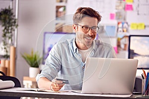 Male Architect In Office Working At Desk Making Online Purchase Using Credit Card On Laptop