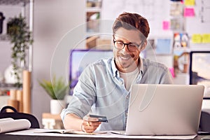 Male Architect In Office Working At Desk Making Online Purchase Using Credit Card On Laptop