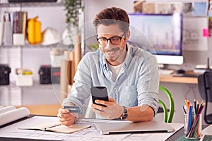 Male Architect In Office Working At Desk Making Online Purchase Using Credit Card On Mobile Phone