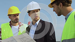 Male architect in suit and safety helmet discussing house scheme with builders