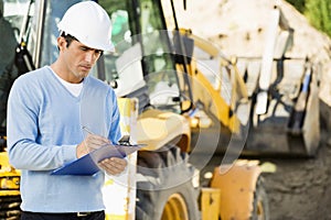 Male architect writing on clipboard against earthmover at construction site