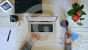 Male arms typing text on laptop keyboard at white wooden desk. Businessman taking pen and making notes on document. Top