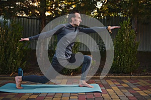 Male athlete exercises outdoors at sunset, assuming warrior pose, stands barefoot on fitness mat. Yoga practice. Asana.
