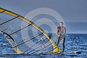 A male athlete is interested in windsurfing. He moves on a Sailboard on a large lake