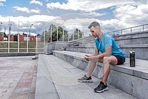 Male athlete sits, holds mobile phone hands, listens music headphones, rest after summer workout in city, background