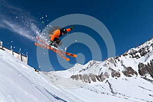 A male athlete skier in an orange trigger makes a jump jump with a grab with flying snow powder against the background