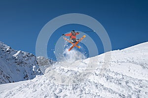 Male athlete skier in an orange trigger makes a jump trick with flying snow powder against the backdrop of snow-capped