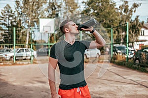 Male athlete, in the summer in the city on the sports ground. In hand a shaker with water and protein. Drinks, resting