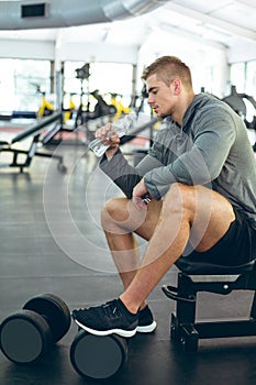 Male athletic drinking water while relaxing on bench in fitness center