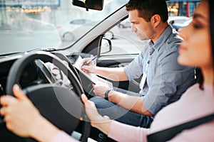 Male auto instructor takes exam in young woman. Model driving car. Hold hands on steering wheel. Male instructor write