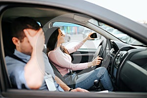 Male auto instructor takes exam in young woman. Shamed guy cover face with hand. Female student driving car and taking