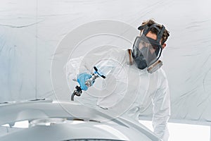 Male auto painter in protective mask and suit painting automobile car bumper in chamber.