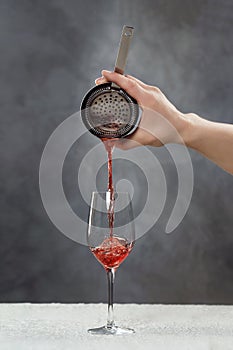 Male bartender is making cocktail pouring alchohol from shaker to wine glass at grey wall background