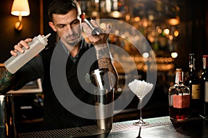 Male bartender pouring a alcoholic drink from the jigger to a steel shaker holding a white plastic bottle