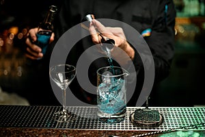 Male bartender pouring a blue alcoholic drink from the jigger to a measuring cup holding a glass bottle