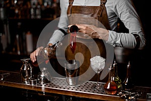 Male bartender pouring a red alcoholic drink from the steel jigger to the cocktail shaker