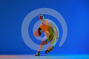 Male basketball player in yellow uniform during game, dribbling ball against blue studio background in neon light.