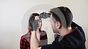 Male beautician in black gloves making permanent makeup procedure on female eyebrows. Beautician cosmetologist in