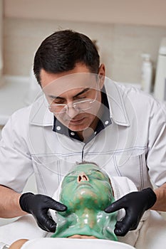 male beautician removing green alginate mask from face of woman