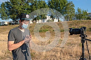 A male blogger in a medical mask in a park filming a video blog on camera. Video blogging, freelance telecommuting