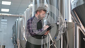 A male brewer in an apron with a beard stands near the beer tanks and writes down the readings. Craft beer production