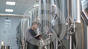 A male brewer in an apron with a beard walks through the beer factory and records the readings on beer tanks. Production