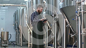 A male brewer with a beard checks the readings of devices on beer tanks using a tablet. Craft beer production