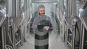 A male brewer with a beard passes by beer tanks and checks the readings using a tablet